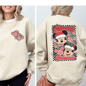 May include: A beige sweatshirt featuring a Christmas-themed design. The front has a small candy cane Mickey Mouse design. The back displays Mickey and Minnie Mouse wearing Santa hats, surrounded by candy canes, with the words "Merry Christmas".