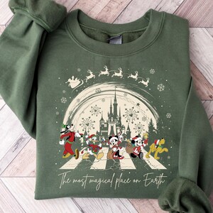 May include: A green sweatshirt with a Christmas-themed design featuring Mickey Mouse and friends walking across a street. The design includes a castle, fireworks, and snowflakes. The text on the sweatshirt reads 'The most magical place on Earth.'