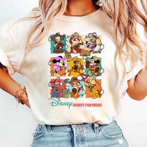 May include: Cream-colored t-shirt featuring a colorful graphic of Disney characters in cowboy attire. The design includes the text "Disney Howdy Partners" in a playful font, with various characters in a grid layout.