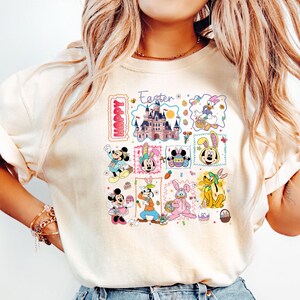 May include: Cream-colored t-shirt featuring a colorful Easter-themed graphic. The design includes Disney characters like Mickey and Minnie Mouse, Donald Duck, and Pluto, along with the text "Happy Easter". The graphic is composed of multiple squares with Easter eggs and a castle.