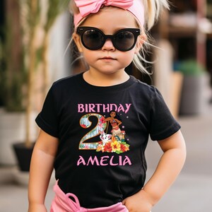 以下が含まれることがあります： ピンクとティール色の「BIRTHDAY 2 AMELIA」の文字が入った黒いTシャツ。シャツには、漫画のキャラクター、花、犬のグラフィックが描かれています。子供はピンクのリボンヘアバンドとサングラスを着用しています。