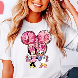 May include: White t-shirt featuring a graphic of Minnie Mouse and Daisy Duck with a pink bow and castle design. The cartoon characters are in a peace sign pose. Pink hearts are in the background.