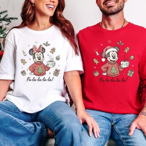 May include: Two Christmas-themed t-shirts, one white and one red, featuring cartoon characters. The white shirt has Minnie Mouse in a red sweater, holding a mug, with the text "Fa-la-la-la-la!". The red shirt has Mickey Mouse in a Santa hat and sweater, also holding a mug, with the same text.