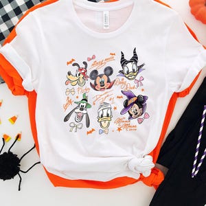Puede incluir: Camiseta blanca con ribete naranja con retratos de personajes de dibujos animados, incluyendo Mickey Mouse, Minnie Mouse y Donald Duck. La camiseta tiene un tema de Halloween con murci&eacute;lagos y calabazas. Tambi&eacute;n se ven pantalones negros y caramelos de Halloween.