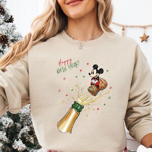May include: Beige sweatshirt with a festive graphic of Mickey Mouse popping a champagne bottle, with the text "Happy New Year!". The design includes stars and a Santa hat. The sweatshirt is paired with red and white plaid pajama bottoms.