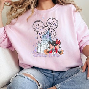 May include: A light pink sweatshirt with a Disney Family design. The graphic features Mickey and Minnie Mouse, a castle, fireworks, and the words "Disney Family" in purple script. The sweatshirt has long sleeves.