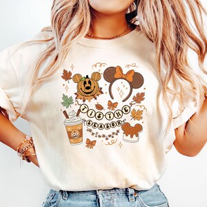 May include: Cream-colored t-shirt featuring a fall-themed graphic. The design includes a pumpkin with a Mickey Mouse face, a Minnie Mouse ear headband, fall leaves, a coffee cup, and the text "Tis the Season."