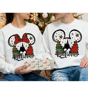 May include: Two white sweatshirts with a Christmas theme. The sweatshirts feature a Mickey Mouse head with a red bow and green and red plaid trees. The text 'Believe' is printed below the image. The sweatshirts are worn by two people.