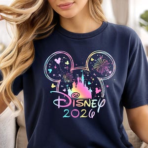 May include: Navy blue t-shirt featuring a colorful Disney design with Mickey Mouse ears, a castle, fireworks, and the text "Disney 2026". The design incorporates pastel colors and a whimsical aesthetic.