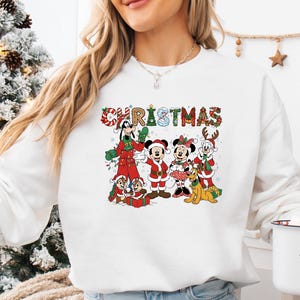 May include: White sweatshirt featuring a colorful Christmas graphic with Disney characters. The word "CHRISTMAS" is displayed above the characters. The graphic includes Mickey Mouse, Minnie Mouse, Goofy, Donald Duck, Chip and Dale, and Pluto in festive attire.