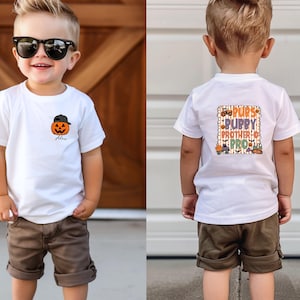 May include: A young child wearing a white t-shirt with a pumpkin graphic and the name "Alex" printed on the front. The back of the shirt has the text "Pups Puppy Brother Pro" in a square design. The child is also wearing brown shorts and white sneakers.