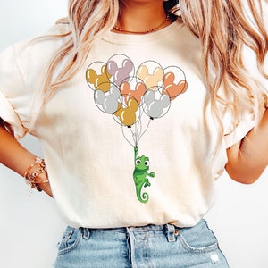 May include: A white t-shirt with a green chameleon holding onto a bunch of colorful balloons with Mickey Mouse ears. The balloons are in shades of gray, pink, yellow, and orange.