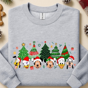 May include: A light gray sweatshirt featuring a festive design with cartoon characters wearing Santa hats and Christmas trees. The design includes Mickey Mouse, Minnie Mouse, Donald Duck, and Goofy. The sweatshirt is a Gildan brand.