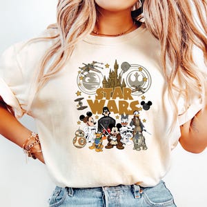 May include: Cream-colored t-shirt featuring a graphic design with the words "STAR WARS" and various characters from the Star Wars and Disney universes. The design includes Mickey Mouse, Darth Vader, and R2-D2.