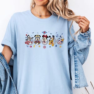 May include: Light blue t-shirt featuring a nautical-themed graphic of Disney characters including Goofy, Mickey, Minnie, Pluto, and Donald Duck. The characters are in circular frames with anchors, stars, and ship wheels.