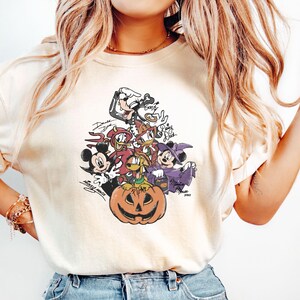 May include: Cream-colored t-shirt featuring a Halloween-themed graphic. The graphic includes various cartoon characters, including Mickey Mouse, Donald Duck, and Goofy, surrounding a carved pumpkin. The design has a vintage, slightly faded appearance.