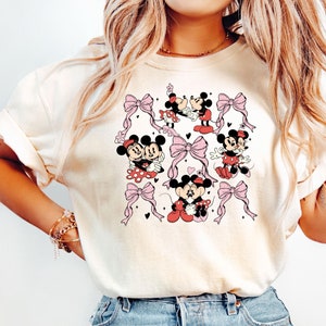 May include: A white t-shirt with a pattern of Mickey Mouse and Minnie Mouse in various poses. The pattern includes pink bows and hearts.