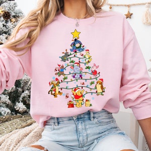 Pu&ograve; includere: Una felpa rosa chiaro con un design ad albero di Natale composto da personaggi di Winnie the Pooh. L'albero &egrave; decorato con luci a stringa e una stella gialla. I personaggi indossano cappelli di Babbo Natale.