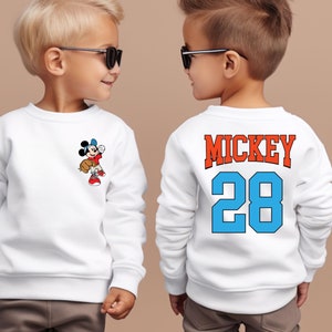 May include: Two children wearing white sweatshirts with a Mickey Mouse design. One sweatshirt has a small Mickey Mouse graphic on the front, while the other has "MICKEY 28" printed on the back in blue and orange.