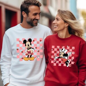 Pode incluir: Um casal usando su&eacute;teres combinando com tema do Dia dos Namorados. O su&eacute;ter do homem &eacute; branco com um padr&atilde;o xadrez rosa e branco e uma imagem de desenho animado do Mickey Mouse segurando uma caixa de chocolates em forma de cora&ccedil;&atilde;o. O su&eacute;ter da mulher &eacute; vermelho com um padr&atilde;o xadrez rosa e branco e uma imagem de desenho animado da Minnie Mouse com asas de anjo segurando um arco e flecha.
