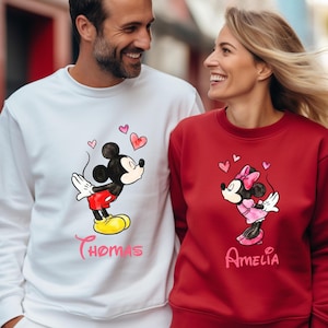 May include: Matching white and red sweatshirts featuring Mickey and Minnie Mouse graphics with hearts. The white sweatshirt has the name "Thomas" and the red sweatshirt has the name "Amelia". The sweatshirts are a couple's matching set.