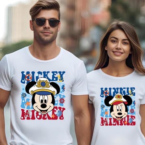May include: White t-shirts with a nautical themed Mickey Mouse and Minnie Mouse design. Mickey Mouse is wearing a captain's hat and Minnie Mouse is wearing a captain's hat with a red bow. The shirts have the text "Mickey Mickey" and "Minnie Minnie" on them.