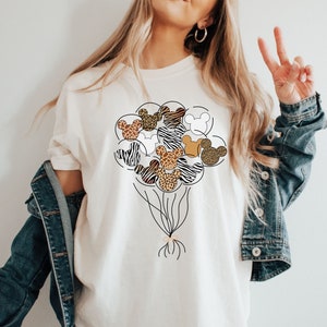Puede incluir: Camiseta blanca con un gr&aacute;fico de un mont&oacute;n de cabezas de Mickey Mouse con estampado de animales en forma de globos.