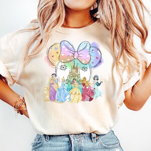 May include: A white t-shirt with a colorful graphic of Disney princesses and Cinderella's castle. The graphic is in the shape of Minnie Mouse ears with a pink bow.
