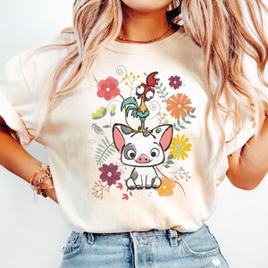 Puede incluir: Una camiseta blanca con una imagen de dibujos animados de un cerdo y un gallo en un dise&ntilde;o floral. El cerdo es blanco con manchas negras y el gallo es rojo, amarillo y verde. Las flores son rosas, amarillas, naranjas y moradas.
