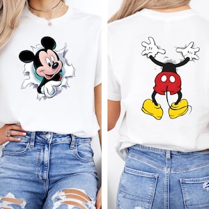 Puede incluir: Camiseta blanca con un dise&ntilde;o de Mickey Mouse. La parte delantera muestra a Mickey rompiendo un efecto rasgado, mientras que la parte trasera muestra la pose ic&oacute;nica de Mickey. La camiseta se combina con unos vaqueros azules.