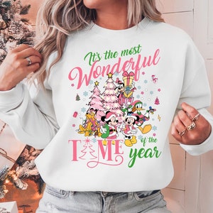 May include: White sweatshirt with a pink and green Christmas design featuring Mickey Mouse and friends. The text on the sweatshirt reads "It's the most Wonderful Time of the Year."