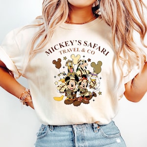 K&ouml;nnte beinhalten: Ein wei&szlig;es T-Shirt mit einem braunen Aufdruck von Mickey Mouse und seinen Freunden auf einem Safari-Abenteuer. Der Text "Mickey's Safari Travel & Co" ist &uuml;ber dem Aufdruck gedruckt.