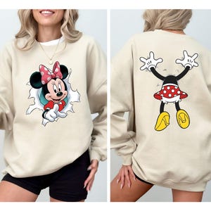 K&ouml;nnte beinhalten: Beiges Sweatshirt mit Minnie Mouse-Motiv. Auf der Vorderseite ist Minnie zu sehen, die aus einem Loch kommt, w&auml;hrend die R&uuml;ckseite sie von hinten zeigt. Minnie tr&auml;gt einen rot-wei&szlig; gepunkteten Rock und eine Schleife mit gelben Schuhen.