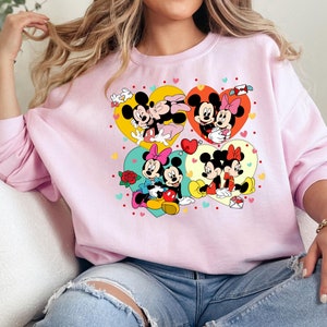 May include: Pink sweatshirt with a graphic print of Mickey Mouse and Minnie Mouse in various poses. The graphic features hearts, flowers, and other romantic details.