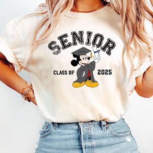 Op de afbeelding: Wit T-shirt met een zwarte afbeelding van Mickey Mouse die een afstudeerpet draagt en een diploma vasthoudt. De tekst "Senior Class of 2025" is onder de afbeelding gedrukt.
