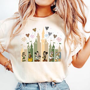 May include: A white t-shirt with a graphic of a castle, fireworks, and Disney characters. The characters are Mickey Mouse, Minnie Mouse, Donald Duck, Goofy, Pluto, and Chip and Dale. The castle is in shades of green and brown. The fireworks are red, yellow, and blue. The hearts are in various colors and patterns.