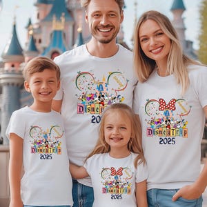 May include: A family of four wearing white t-shirts with a Disney Trip 2025 design. The shirts feature Mickey Mouse ears with a red bow on the women's shirt and a blue bow on the children's shirts. The shirts also feature a colorful illustration of Mickey Mouse and other Disney characters.