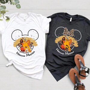 Puede incluir: Dos camisetas con un dise&ntilde;o de cabeza de Mickey Mouse en blanco y negro con los personajes de El Rey Le&oacute;n Simba, Nala, Tim&oacute;n y Pumba. El texto "Hakuna Matata" est&aacute; impreso debajo del dise&ntilde;o. Una camiseta es blanca y la otra es negra.