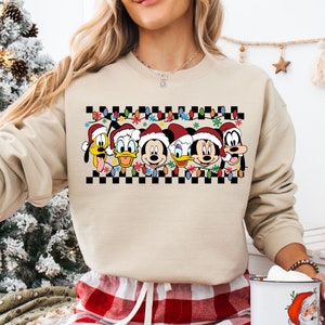 May include: A beige sweatshirt with a checkered pattern and a row of Disney characters wearing Santa hats. The characters include Mickey Mouse, Minnie Mouse, Donald Duck, Goofy, and Pluto. The characters are surrounded by Christmas lights.