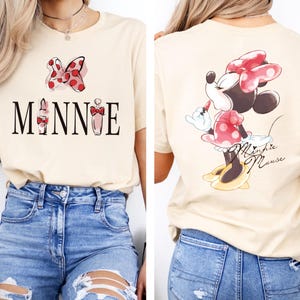 May include: Beige t-shirt featuring a Minnie Mouse design. The front of the shirt displays the word "MINNIE" with a bow and lipstick graphic. The back shows Minnie Mouse in a red polka dot dress and bow. The shirt is paired with blue jeans.