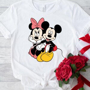 May include: White t-shirt with a graphic of Mickey Mouse and Minnie Mouse holding hands. The graphic is in black, red, and yellow.