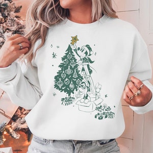 May include: White sweatshirt with a green illustration of Mickey Mouse and Pluto decorating a Christmas tree. Mickey is on a ladder placing a star on top of the tree. Pluto is sitting in a box with presents.