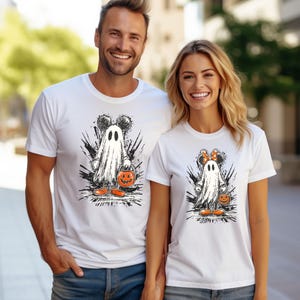 Peut inclure: T-shirts blancs avec un motif d'Halloween. Le motif repr&eacute;sente un fant&ocirc;me avec des oreilles de Mickey Mouse tenant une citrouille. Le fant&ocirc;me est entour&eacute; de coups de pinceau noirs. Le t-shirt de la femme a un n&oelig;ud Minnie Mouse.