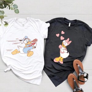 May include: A white t-shirt with a cartoon image of Donald Duck running with a bouquet of flowers. A black t-shirt with a cartoon image of Daisy Duck with hearts floating above her head.