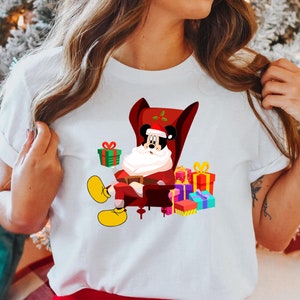 Op de afbeelding: Wit T-shirt met een cartoon van Mickey Mouse als kerstman, zittend in een rode fauteuil. Mickey is omringd door kleurrijk verpakte cadeaus. Het ontwerp bevat een kerstmuts en gele schoenen.