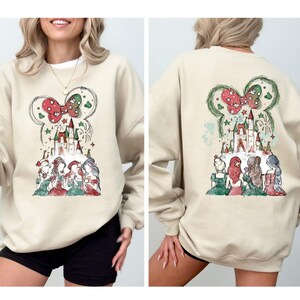 May include: Beige sweatshirt featuring a whimsical design of a castle, bow, and characters. The front and back of the sweatshirt showcase the same design. The design includes red, green, and white accents, creating a festive and charming aesthetic.