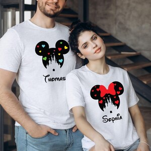 May include: A matching pair of white t-shirts for a couple, featuring black silhouettes of Mickey Mouse and Minnie Mouse heads with a castle inside. The Mickey Mouse shirt has colourful hearts and the text "Thomas" below. The Minnie Mouse shirt has red hearts and a red bow and the text "Sophia" below.