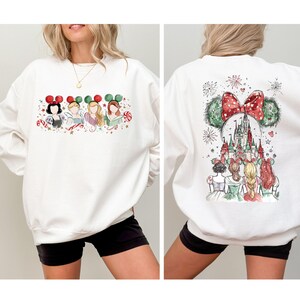 May include: White sweatshirt featuring a watercolor design of Disney princesses with holiday-themed Mickey Mouse ears on the front. The back of the sweatshirt showcases a castle and more princesses with fireworks.