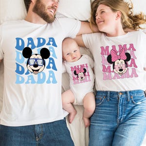 May include: A family of three wearing matching white t-shirts with a Mickey Mouse and Minnie Mouse design. The shirts say "Dada", "Mama", and "Mini" in a retro font.