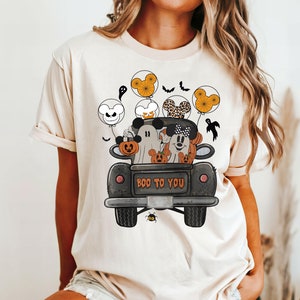 Peut inclure: Un t-shirt blanc avec une illustration noire et blanche de Mickey Mouse, Minnie Mouse et un fant&ocirc;me dans un pick-up noir avec des citrouilles. Le camion porte l'inscription "BOO TO YOU" &agrave; l'arri&egrave;re. Les personnages tiennent des ballons avec des oreilles de Mickey Mouse.